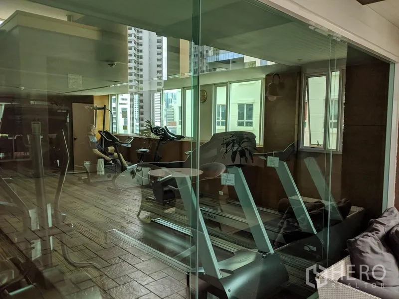 Condominium for rent in Watthana, Bangkok - Fitness room with treadmills and weight machines behind glass, overlooking city buildings.