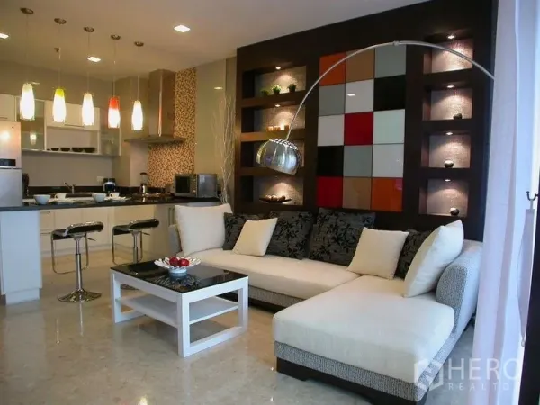 Condominium for rent in Khlong Luang, Pathum Thani - Open-plan living area with L-shaped sofa, coffee table and kitchen lit by colorful pendant lights.