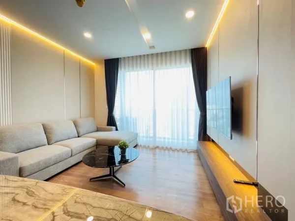 Condominium for rent in Khlong Toei, Bangkok - Bright living room with an L-shaped grey sofa, floor-to-ceiling windows, warm LED lighting and a wall-mounted TV in a modern Bangkok condo near BTS Thonglor.