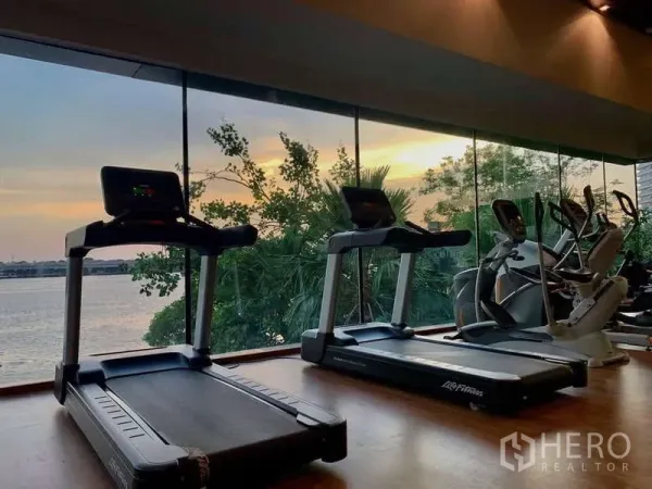 Condominium for rent or sale in Bangkok - Riverside fitness room with treadmills facing floor-to-ceiling windows at sunset.