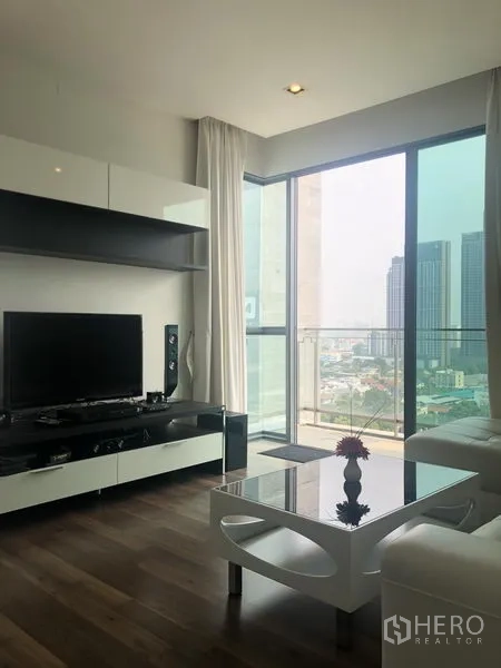 Condominium for rent or sale in Phra Khanong, Bangkok - Living area with TV console and floor-to-ceiling windows opening to a balcony with city skyline near BTS Punnawithi.
