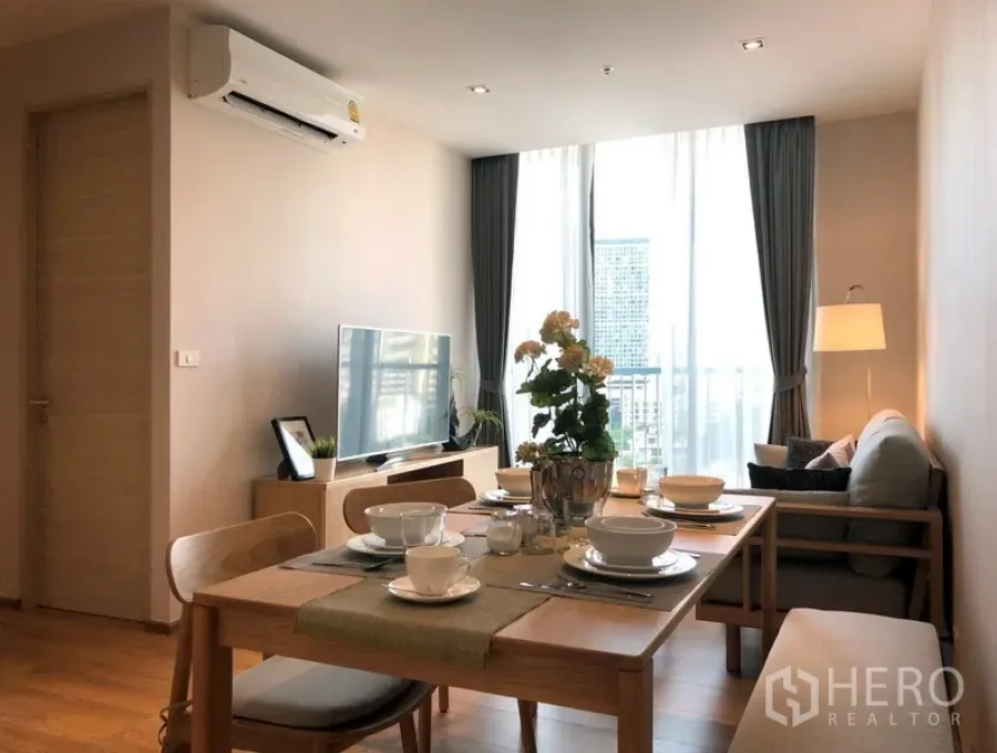 Condominium for sale in Khlong Toei, Bangkok - Bright living and dining area with a wooden table set, sofa, TV console and floor-to-ceiling windows showing a city view.