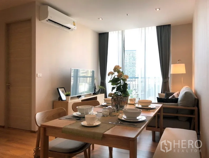 Condominium for sale in Khlong Toei, Bangkok - Bright living and dining area with a wooden table set, sofa, TV console and floor-to-ceiling windows showing a city view.