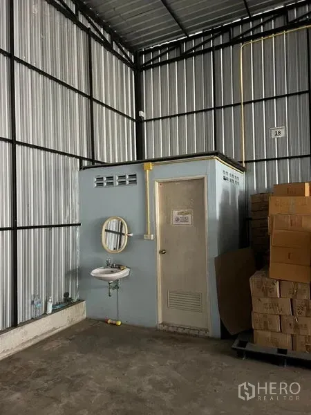 货栈 出租在Lat Phrao, Bangkok - 仓库一角设有独立卫生间与洗手盆，管线外露，旁边堆有纸箱货物。