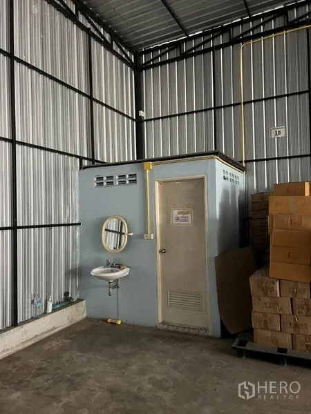 货栈 出租在Lat Phrao, Bangkok - 仓库一角设有独立卫生间与洗手盆，管线外露，旁边堆有纸箱货物。