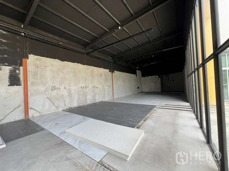 Retail Space for rent in Bang Na, Bangkok - Open-plan retail interior with high black ceiling, unfinished walls, and mixed tile flooring beside a full-height glass storefront.