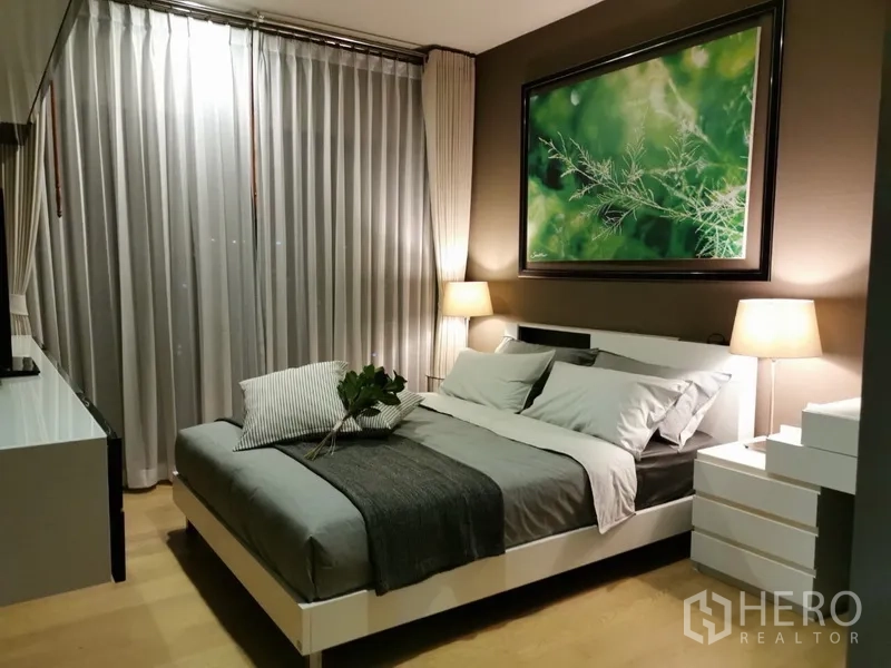 公寓 出售在Bangkok - 主卧配大床、床头灯、自然主题装饰画与转角窗帘。