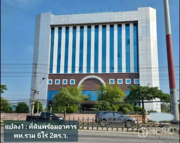独立商业地产 出租在Krathum Baen, Samut Sakhon - 带蓝色竖向玻璃的现代中高层建筑街景，前方有行道树与车辆。