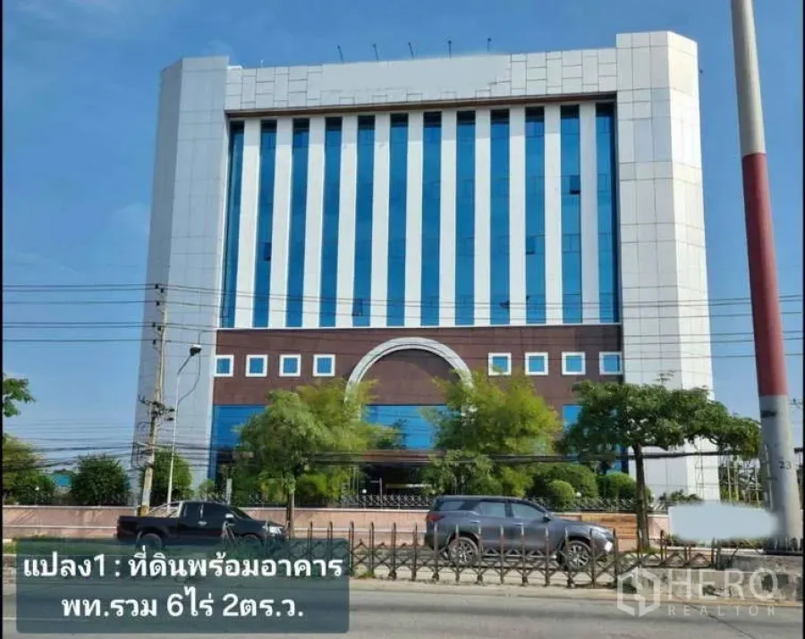 Standalone for rent in Krathum Baen, Samut Sakhon - Street view of a modern mid‑rise commercial building with blue vertical glass panels, trees, and cars in front.