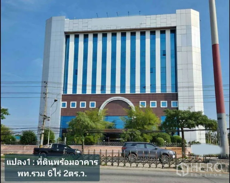 Standalone for rent in Krathum Baen, Samut Sakhon - Street view of a modern mid‑rise commercial building with blue vertical glass panels, trees, and cars in front.