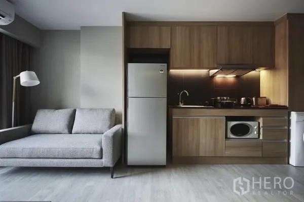 Apartment for rent in Khlong Toei, Bangkok - Open-plan living area with gray sofa beside a full kitchen featuring wood cabinets, stainless fridge and microwave.