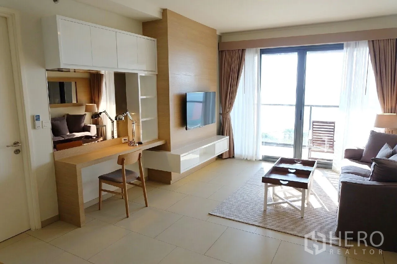 Condominium for rent or sale in Bang Lamung, Chon Buri - Living room with large windows, sofa set, TV wall and a work desk opening to the balcony.