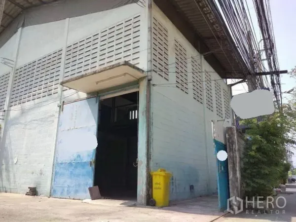 Warehouse for rent in Mueang Nonthaburi, Nonthaburi - Corner warehouse facade with tall sliding door under a small canopy and utility lines along the street.