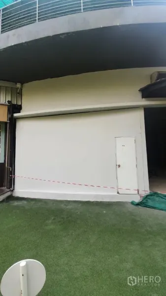 Retail Space for rent in Khlong Toei, Bangkok - Curved shopfront with white roller shutter and side access door opening to an artificial grass forecourt.