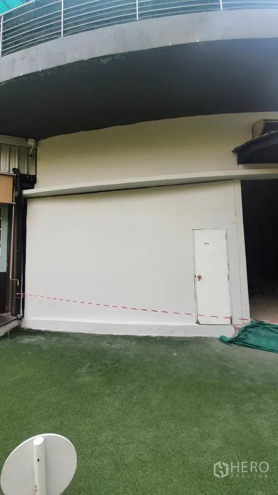 Retail Space for rent in Khlong Toei, Bangkok - Curved shopfront with white roller shutter and side access door opening to an artificial grass forecourt.