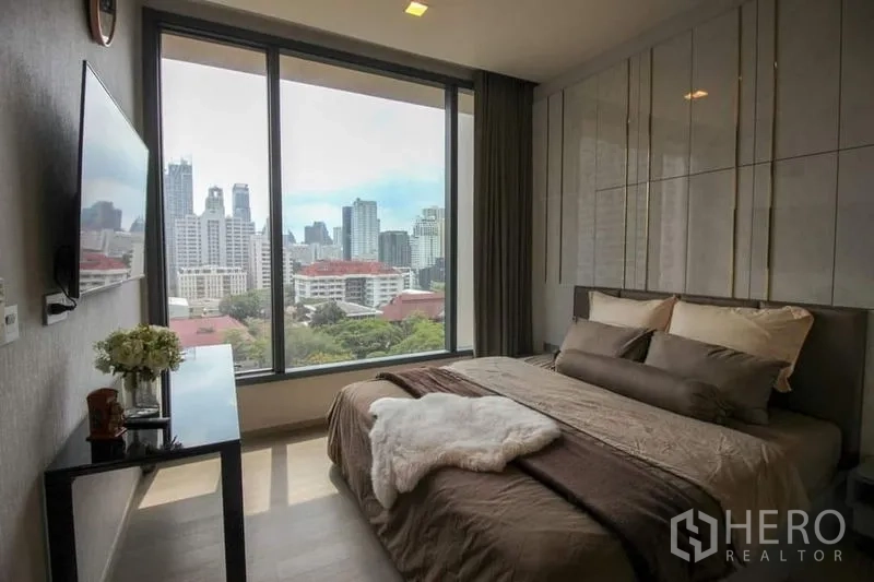Condominium for sale in Watthana, Bangkok - Bedroom with large picture windows showcasing the Sukhumvit city skyline and a TV opposite the bed.