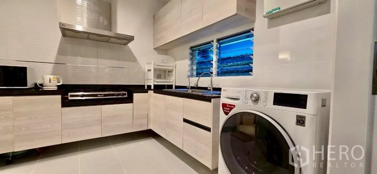 Detached House for rent in Samut Prakan - Modern Thai kitchen with L-shaped cabinets, gas hob, hood and a washer by the window.