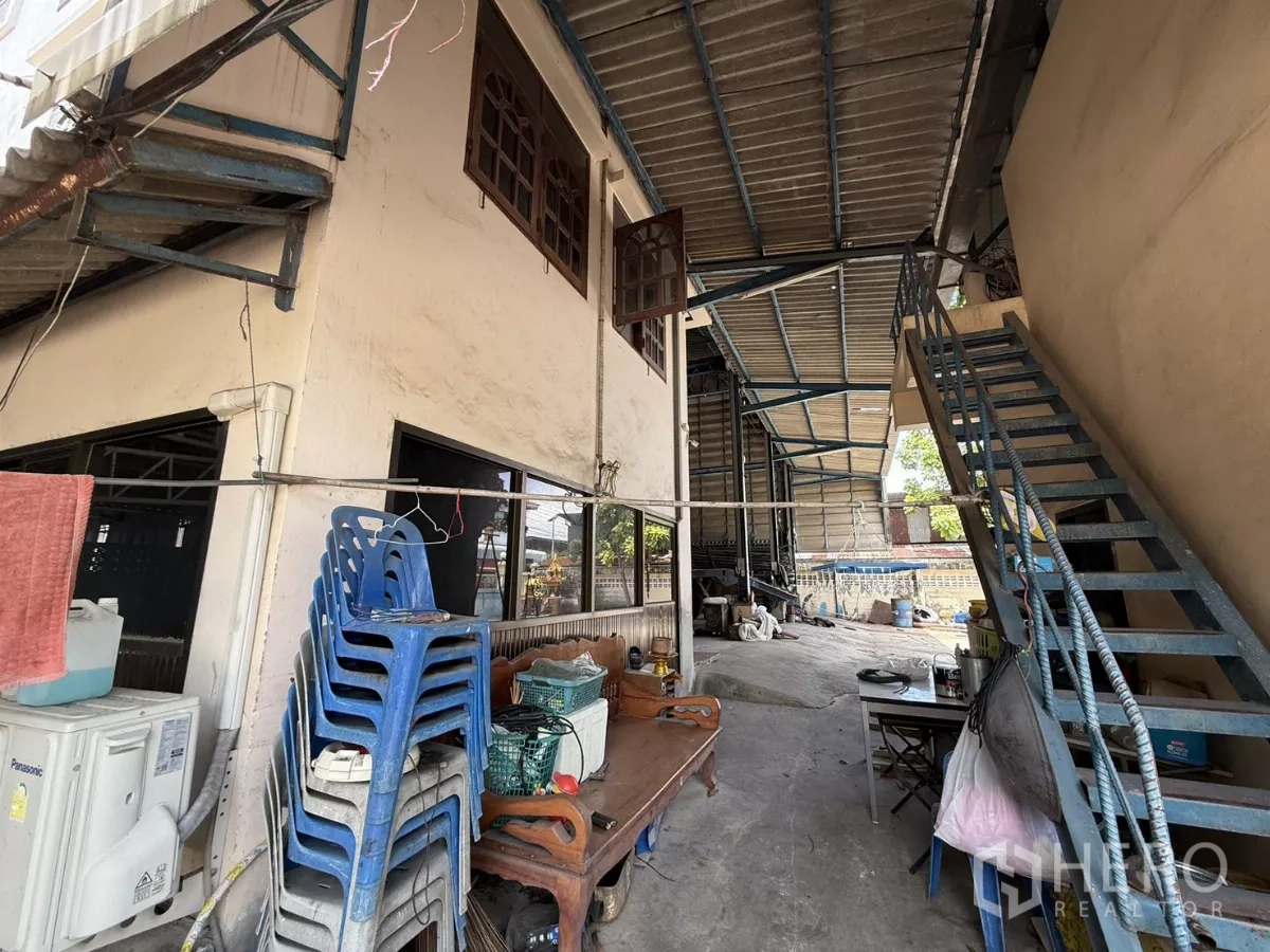 Warehouse for sale in Phra Samut Chedi, Samut Prakan - Covered walkway and metal stairs beside storage area with stacked plastic chairs.