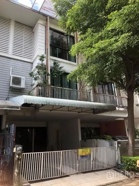 ทาวน์เฮาส์ ขาย - ด้านหน้าทาวน์เฮ้าส์สูง 3 ชั้น มีกำแพงรั้วสีขาว โรงจอดรถมีหลังคา ระเบียง และต้นไม้ใหญ่ให้ร่มเงา