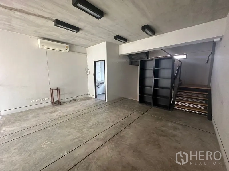 Home Office for rent in Phra Khanong, Bangkok - Open concrete-floored room with black shelving, staircase and a doorway to a bathroom.