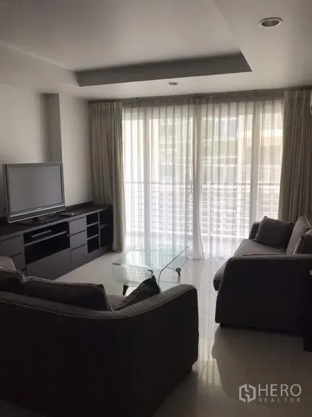 Condominium for rent in Watthana, Bangkok - Bright living room with floor-to-ceiling curtains, TV console, sofa set and glass coffee table opening to a balcony.