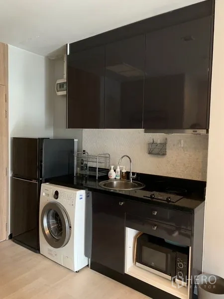 Condominium for sale in Watthana, Bangkok - Modern kitchenette with glossy cabinets, sink, cooktop, microwave, refrigerator, and washing machine.