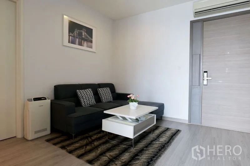 คอนโด ให้เช่าในWatthana, Bangkok - มุมนั่งเล่นอบอุ่น โซฟาดำพรมปูพื้น โต๊ะกลางยกได้ และแอร์ใกล้ประตู