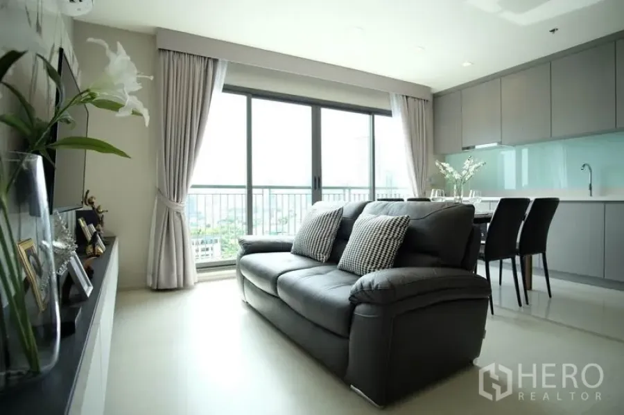 Condominium for rent in Khlong Toei, Bangkok - Bright open-plan living room with leather sofa, dining set, grey built-in kitchen and floor-to-ceiling windows opening to a balcony with city views.