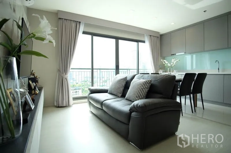 Condominium for rent in Khlong Toei, Bangkok - Bright open-plan living room with leather sofa, dining set, grey built-in kitchen and floor-to-ceiling windows opening to a balcony with city views.