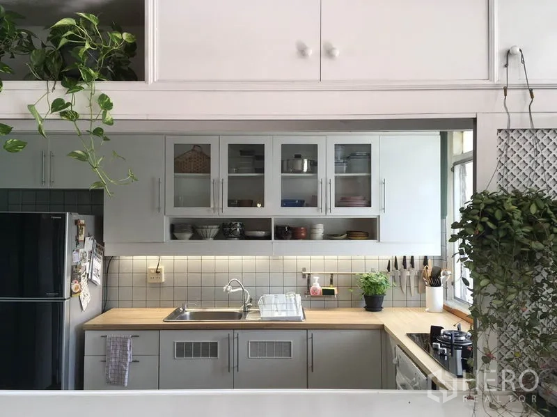 Apartment for rent in Khlong Toei, Bangkok - U-shaped kitchen with white cabinetry, tiled backsplash, wooden countertops, sink, and stovetop, accented by plants.