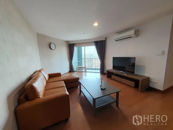 Condominium for rent in Huai Khwang, Bangkok - Living room with L-shaped leather sofa, flat-screen TV and sliding doors to a city-view balcony.