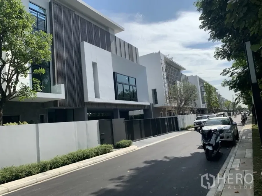 Detached House for rent in Saphan Sung, Bangkok - Quiet estate street lined with modern three-storey houses and roadside parking under trees.