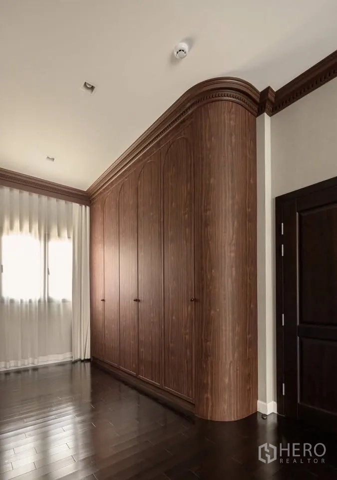 Detached House for rent or sale in Lat Krabang, Bangkok - Curved-corner wood wardrobe beside a window with sheer drapes on dark floors.