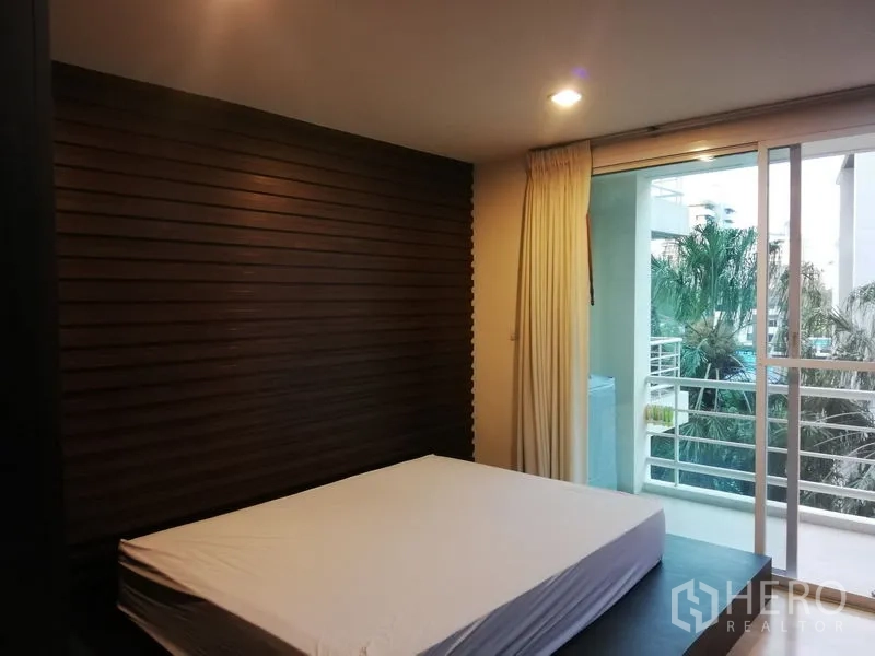 Condominium for rent in Watthana, Bangkok - Master bedroom with wood accent wall and sliding doors to a balcony facing trees and the pool area.