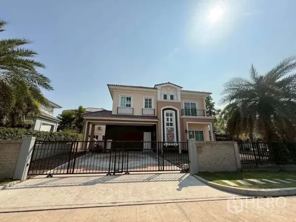 独立式住宅 出售在Bang Khen, Bangkok - 两层独栋别墅，带前院与电动大门车道，棕榈树环绕，天空晴朗。