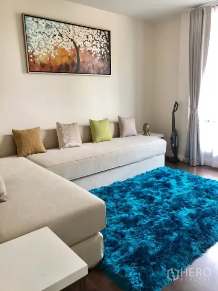 Detached House for rent in Bang Kapi, Bangkok - Cozy family room with long beige sofa, teal shag rug, wall art and a large curtained window.