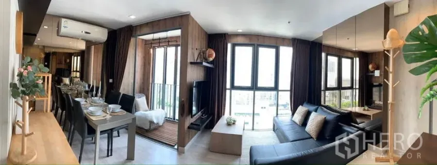 Condominium for rent or sale in Phra Khanong, Bangkok - Open-plan living and dining area with black sectional sofa, mirrored wall, balcony nook and floor-to-ceiling windows.