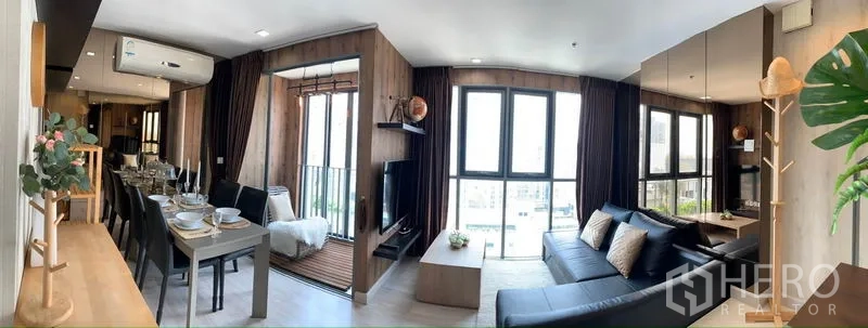 Condominium for rent or sale in Phra Khanong, Bangkok - Open-plan living and dining area with black sectional sofa, mirrored wall, balcony nook and floor-to-ceiling windows.