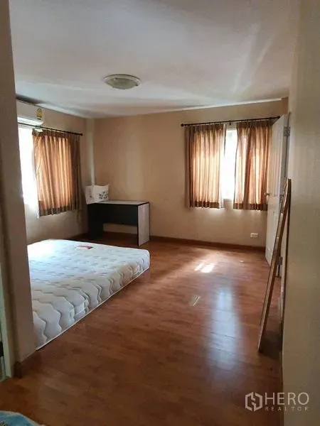 Townhouse for rent in Bueng Kum, Bangkok - Bedroom with wood flooring, two windows with brown curtains, a mattress, and a compact desk.