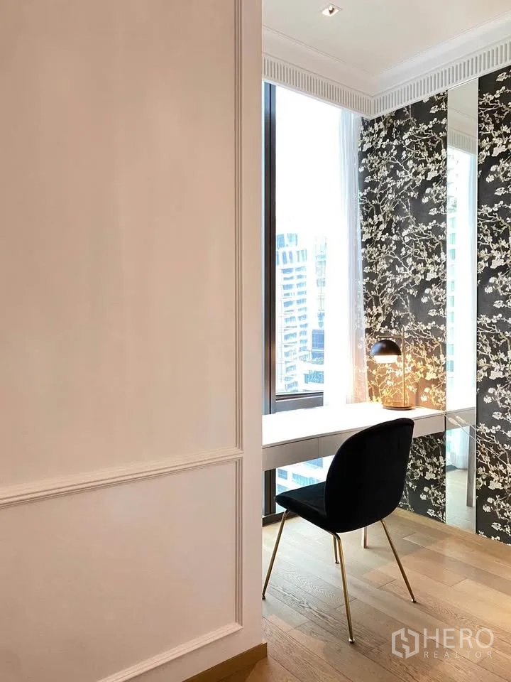 Condominium for rent in Pathum Wan, Bangkok - Bright study nook with built-in white desk, black chair, table lamp, and city view by tall windows and floral wall panels.
