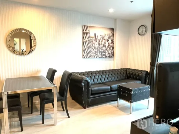 Condominium for sale in Sathon, Bangkok - Living and dining area with a black leather Chesterfield sofa, glass dining table, trunk-style coffee table and curtained window.