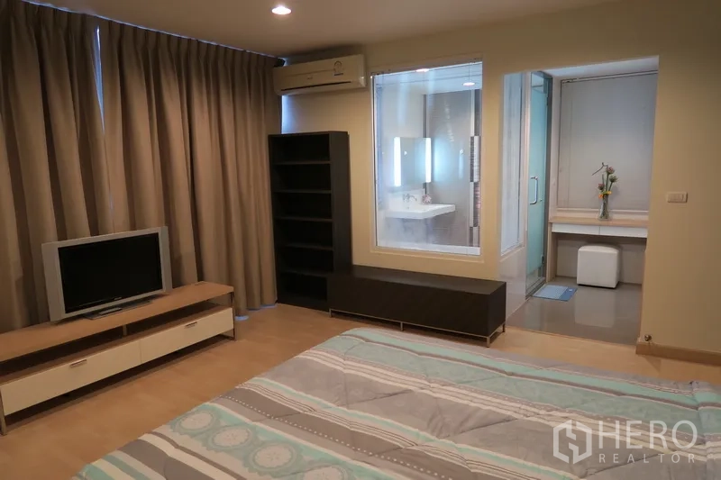 Condominium for rent in Huai Khwang, Bangkok - Master suite with TV stand, bookshelf, air conditioner and glass‑partitioned ensuite bathroom.