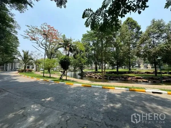 Land for sale in Khlong Luang, Pathum Thani - Landscaped community park with a pond, rocks and mature trees beside a paved road.