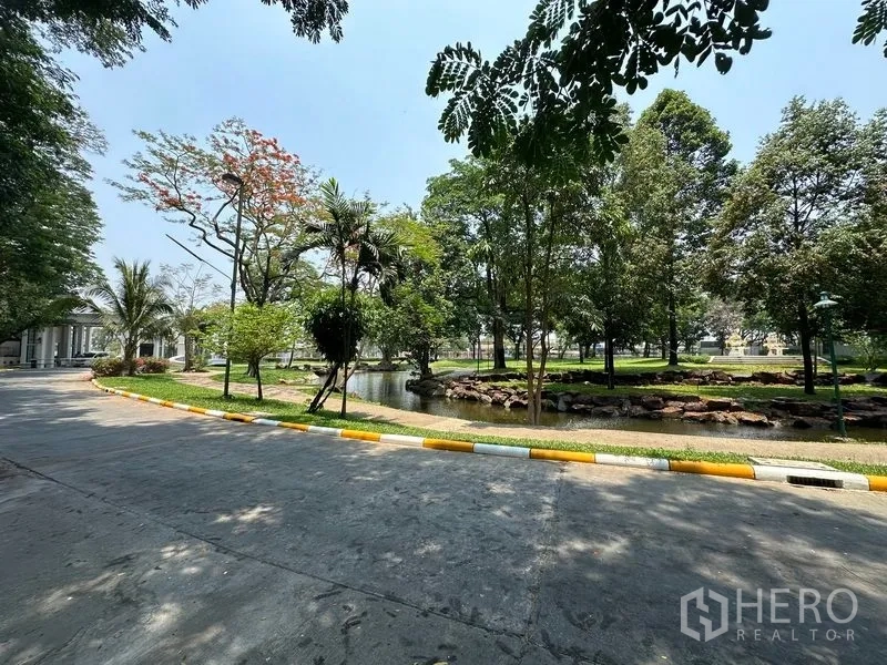 Land for sale in Khlong Luang, Pathum Thani - Landscaped community park with a pond, rocks and mature trees beside a paved road.