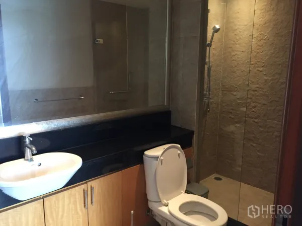 Condominium for rent in Sathon, Bangkok - Modern bathroom with glass-enclosed shower, stone-textured wall, long vanity with vessel sink, and toilet.