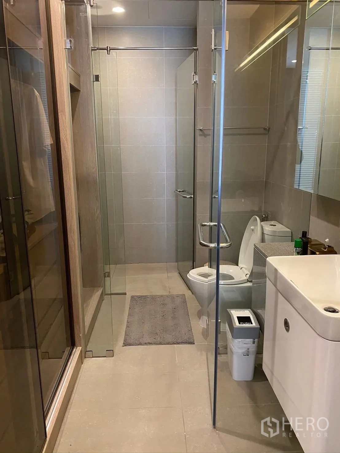Condominium for rent in Pathum Wan, Bangkok - Modern bathroom with glass-enclosed shower, toilet, and vanity in neutral gray tiles.