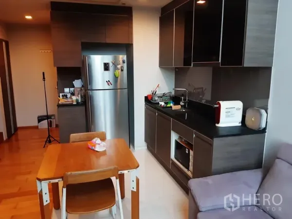 Condominium for rent or sale in Khlong Toei, Bangkok - Modern kitchen with dark cabinetry, stainless refrigerator, storage and a dining table beside it.