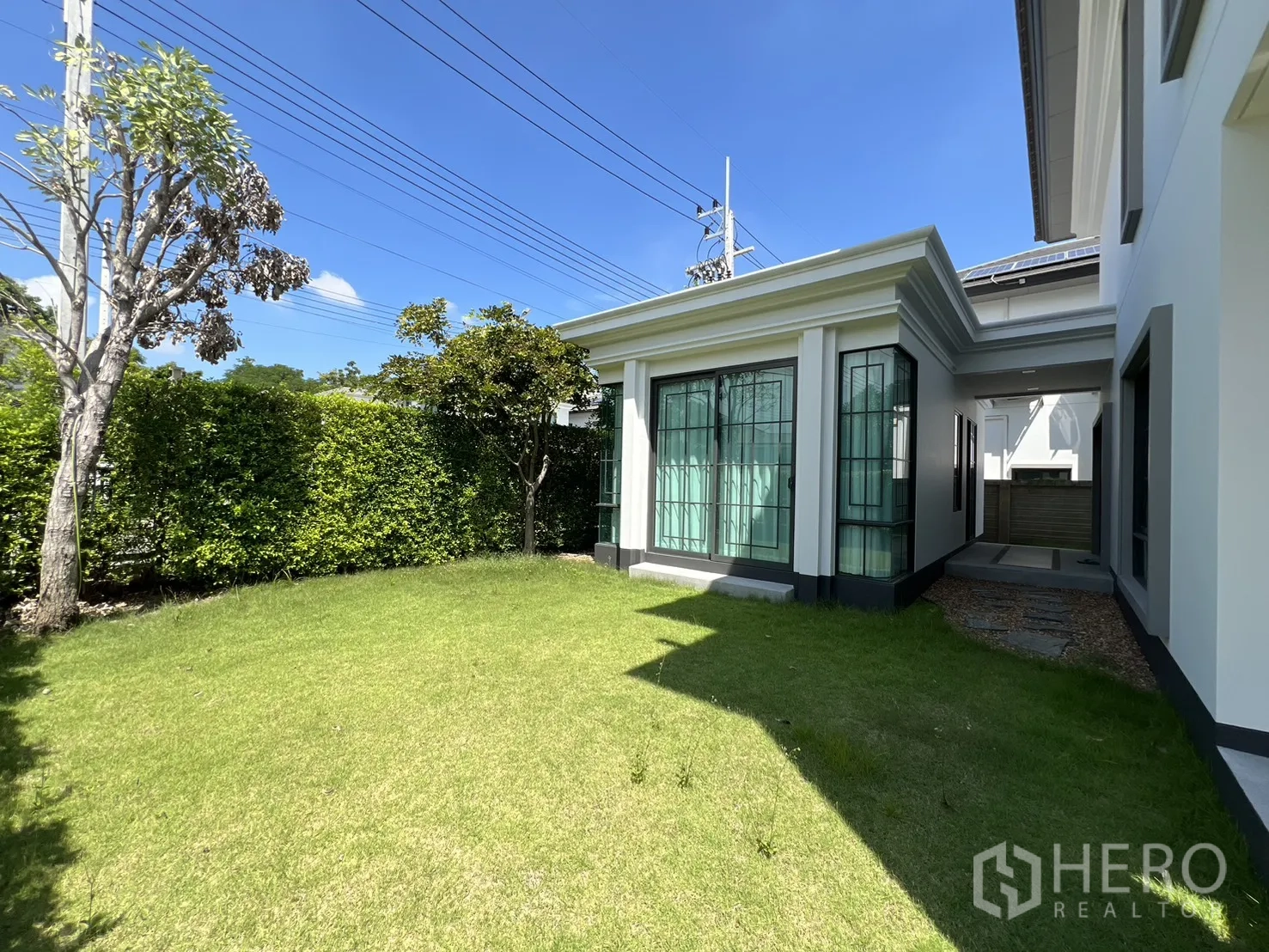 Detached House for rent or sale in Mueang Pathum Thani, Pathum Thani - Backyard lawn bordered by hedges and a bright glass pavilion room.
