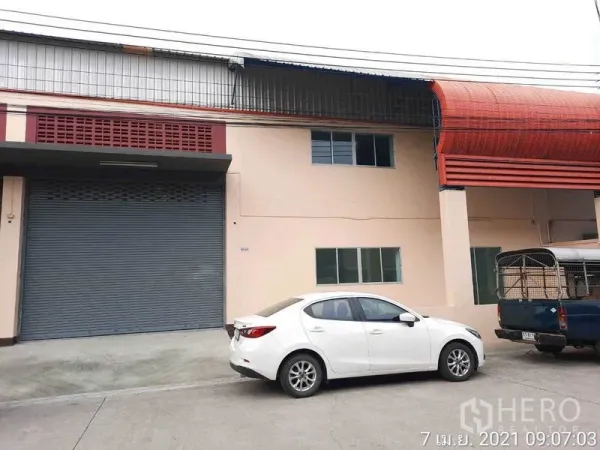 工厂 出租在Bang Bua Thong, Nonthaburi - 工厂外立面，配大尺寸卷帘门、红色雨棚及停车位。