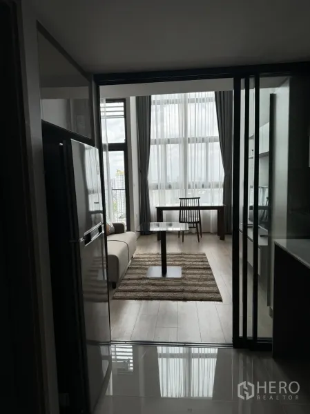 Condominium for rent in Huai Khwang, Bangkok - Modern condo living area with floor-to-ceiling windows, sofa, rug and dining table viewed from the kitchen.